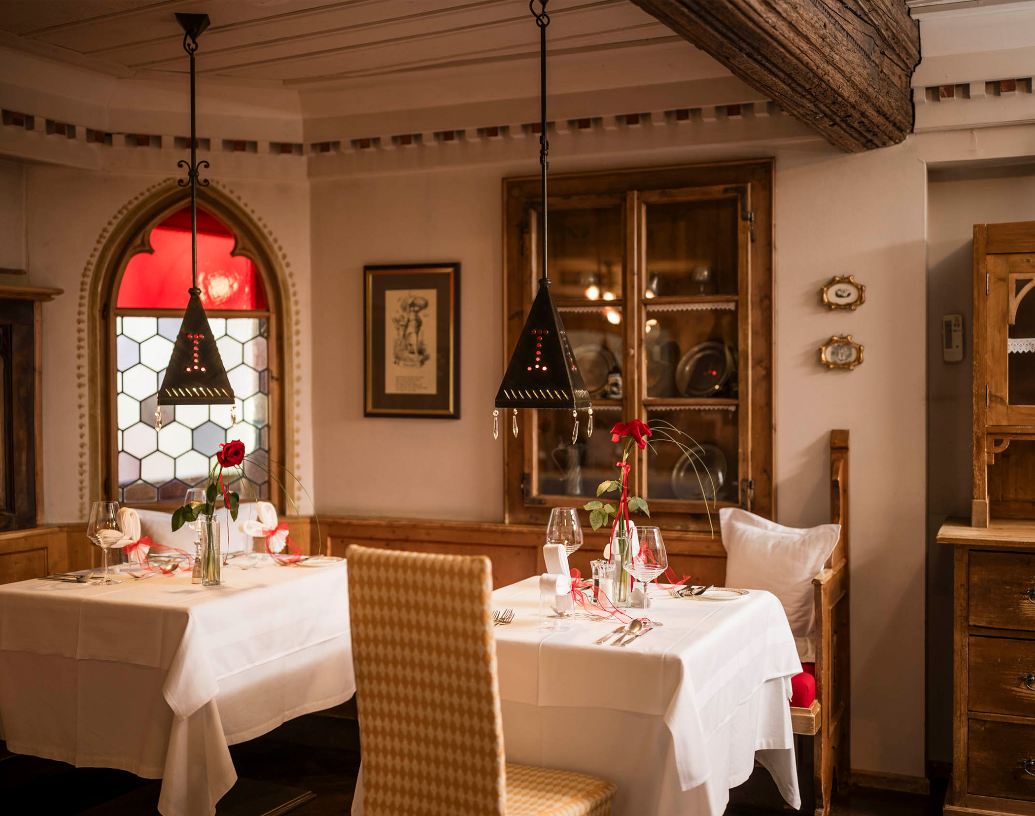 Zwei Tische mit weißen Tischdecken, roten Rosen und eleganten Gedecken in einem gemütlichen, warm beleuchteten Restaurant. - Mirabell Dolomites Hotel