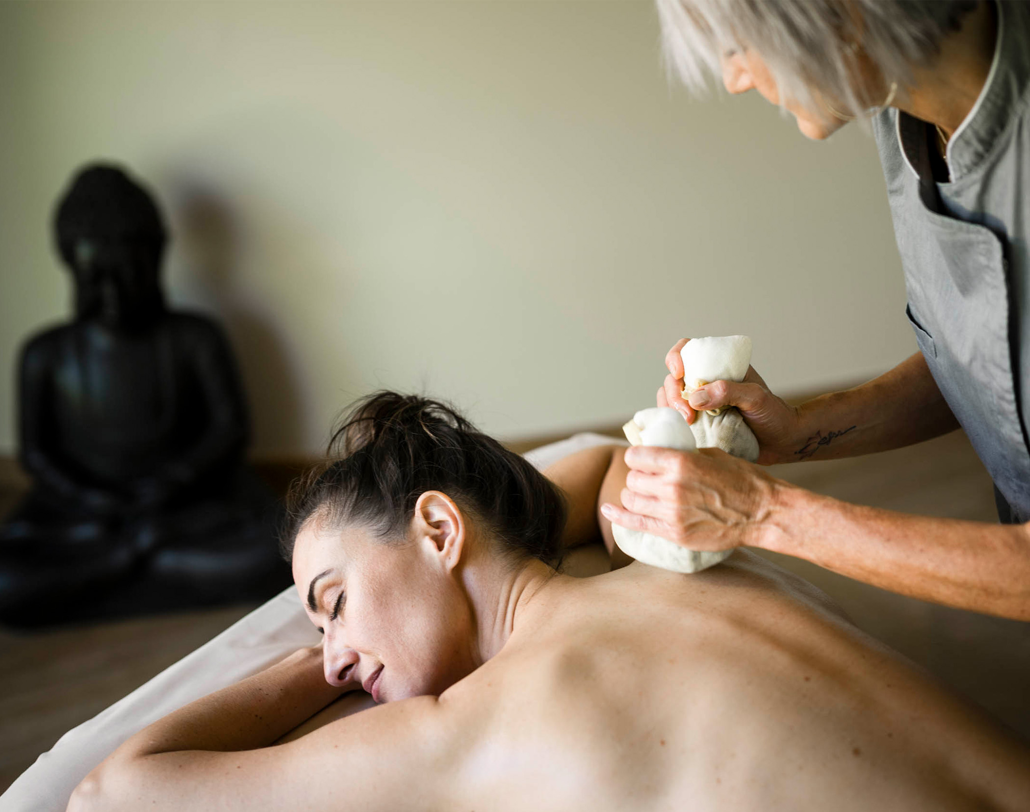 Eine Frau, die in einem Spa eine Kräutermassage am Rücken erhält, im Hintergrund ist eine Buddha-Statue zu sehen. - Mirabell Dolomites Hotel