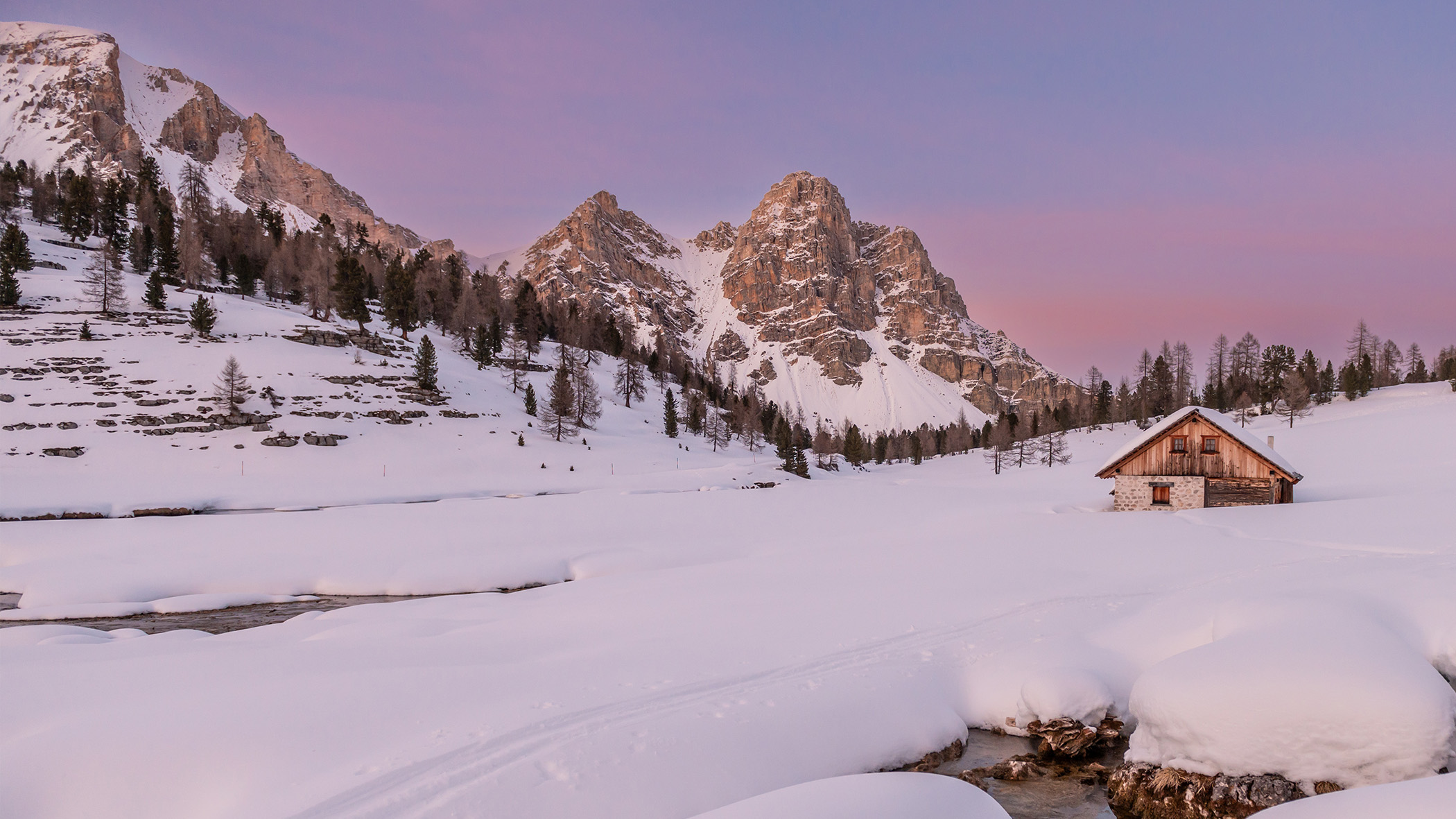 Verschneite Berglandschaft bei Sonnenuntergang mit einer Holzhütte und Bäumen unter einem violett-rosa Himmel. - Mirabell Dolomites Hotel