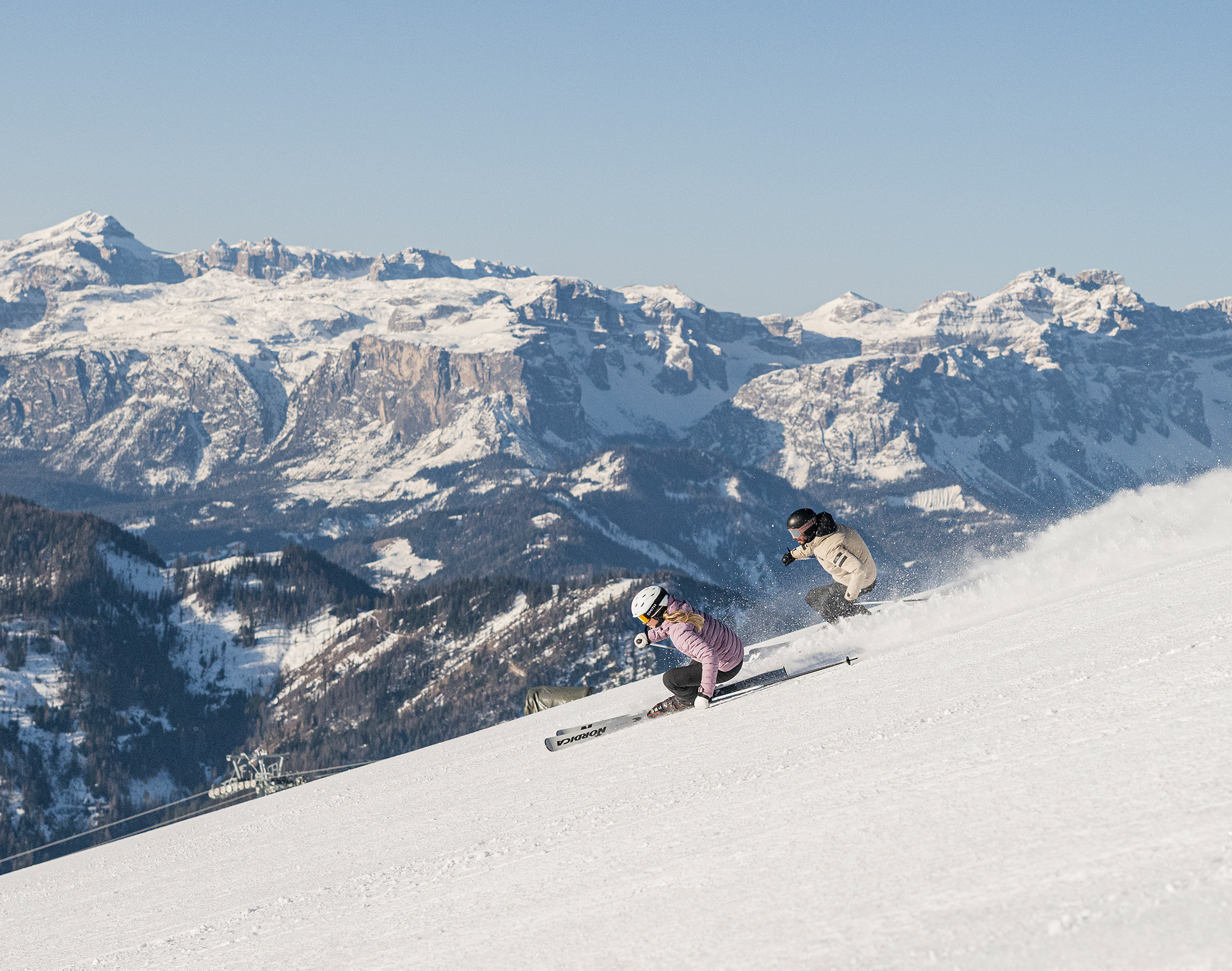 Zwei Personen fahren auf einem verschneiten Hang Ski, im Hintergrund sind Berge und ein klarer Himmel zu sehen. - Mirabell Dolomites Hotel