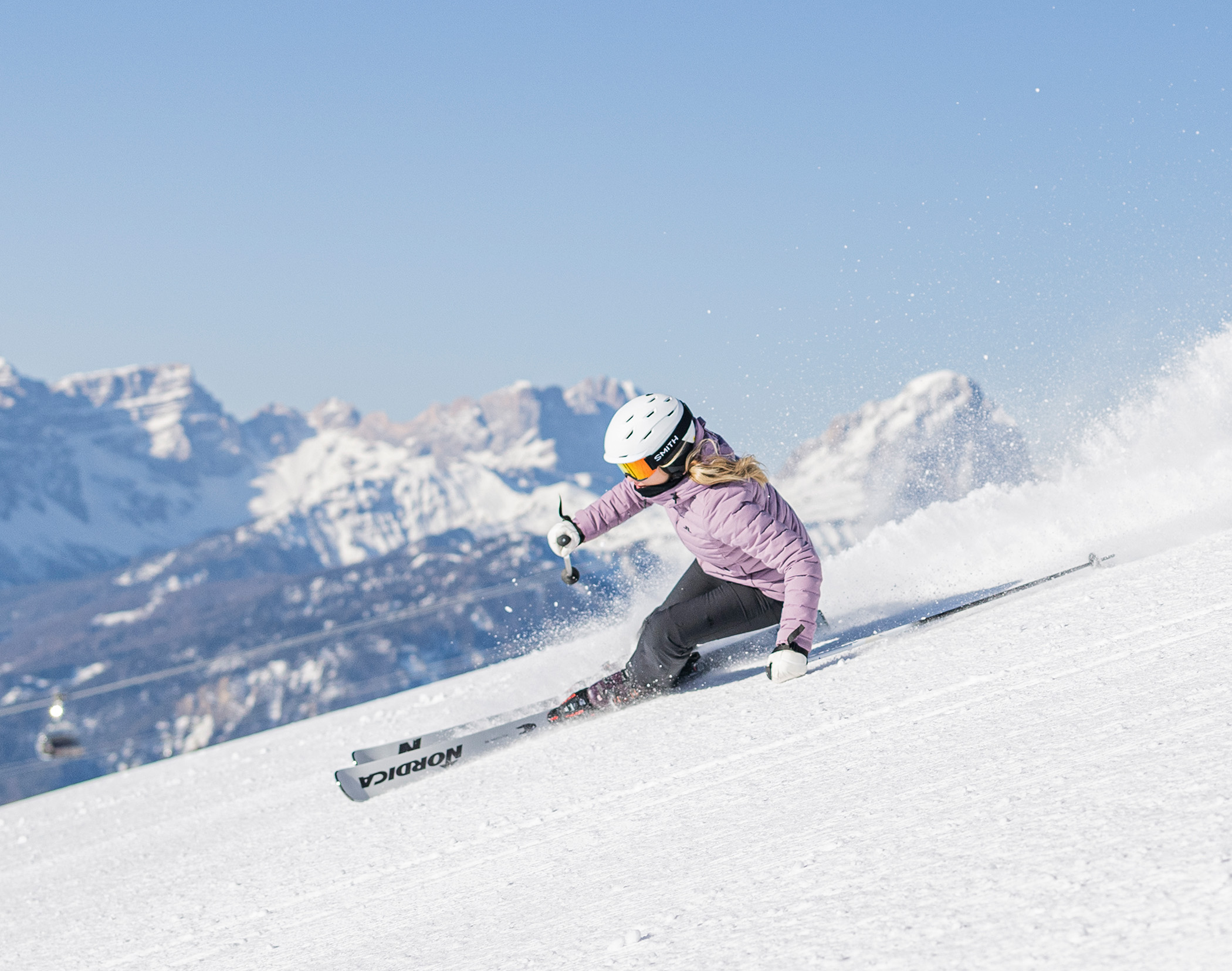 Ein Skifahrer in einer lila Jacke und mit Helm, der auf einem verschneiten Hang eine scharfe Kurve fährt, im Hintergrund sind Berge zu sehen. - Mirabell Dolomites Hotel