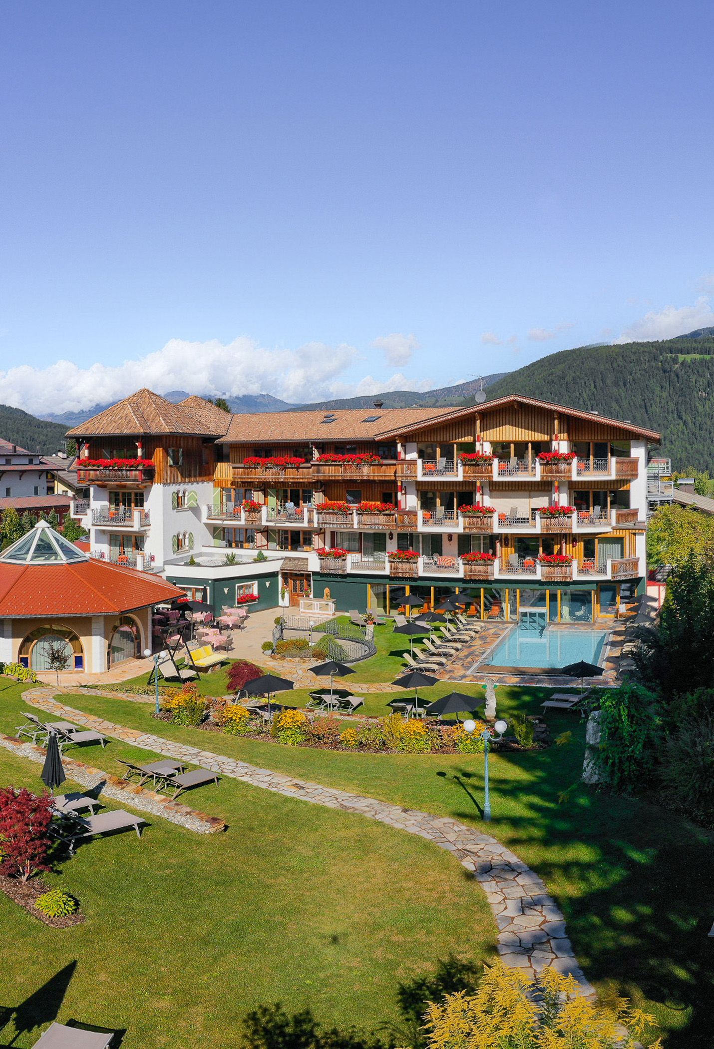 Ein mehrstöckiges Hotel mit blumengeschmückten Balkonen, einem Pool und einem Garten in den Bergen. - Mirabell Dolomites Hotel