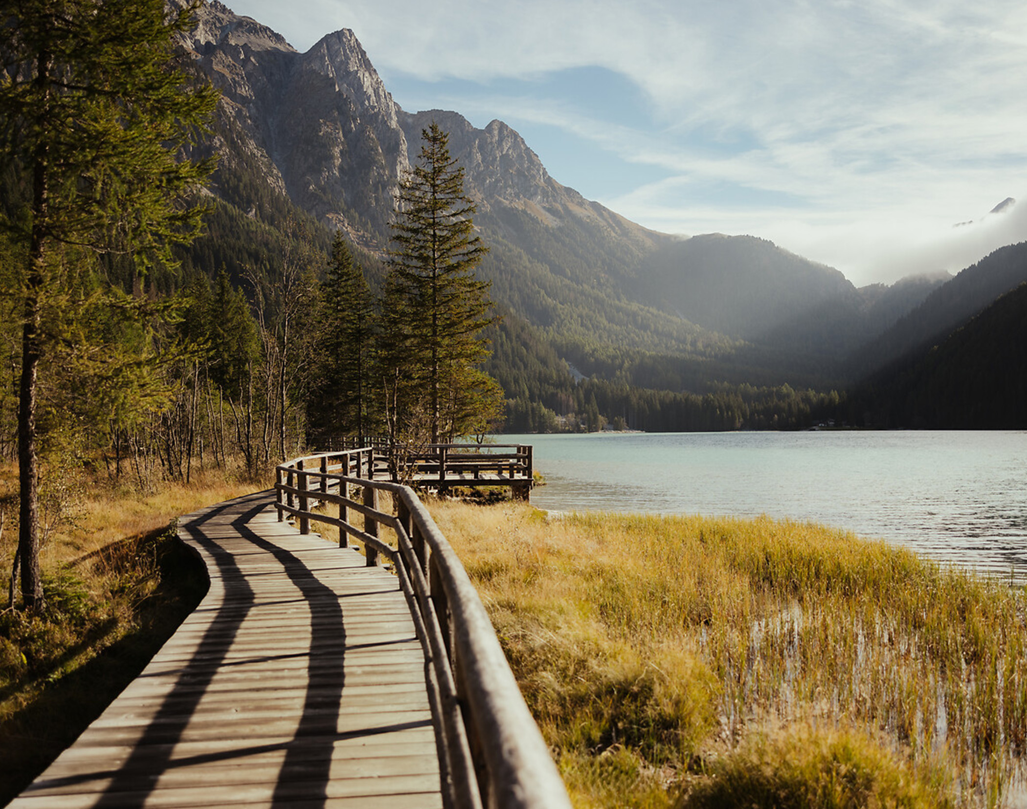 Wooden boardwalk beside a calm lake, mountains and trees in the background under a clear sky. - Mirabell Dolomites Hotel