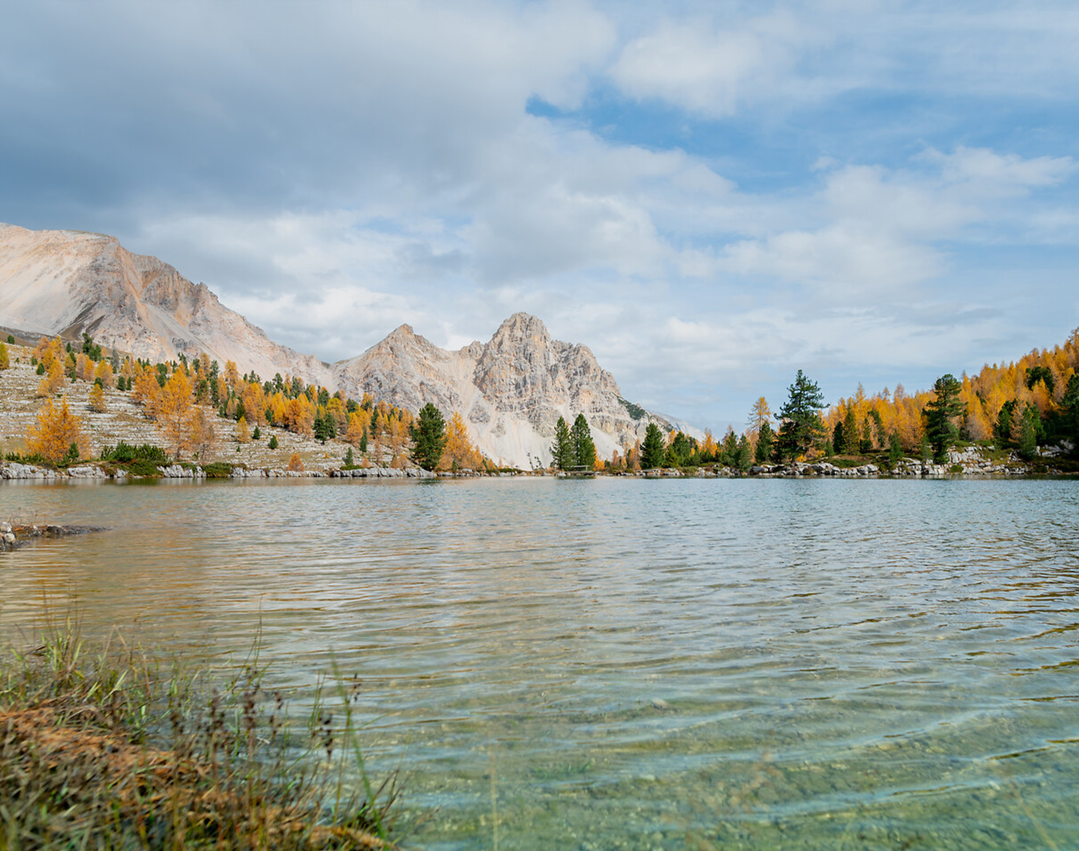 Clear lake with ripples, autumn trees, and rocky mountains under a partly cloudy sky in the background. - Mirabell Dolomites Hotel