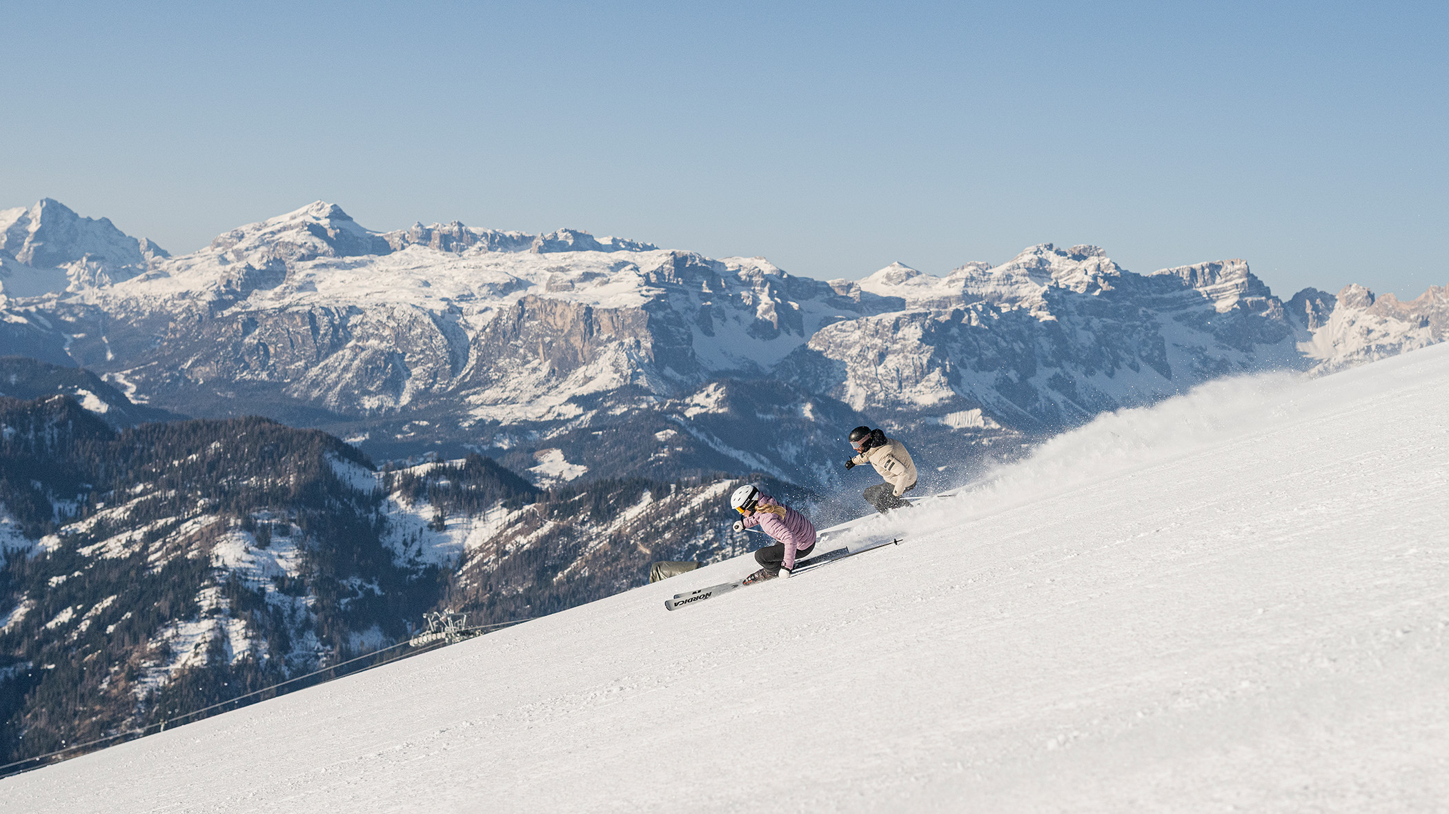 Zwei Personen fahren auf einem verschneiten Hang unter klarem Himmel mit Bergen im Hintergrund Ski. - Mirabell Dolomites Hotel