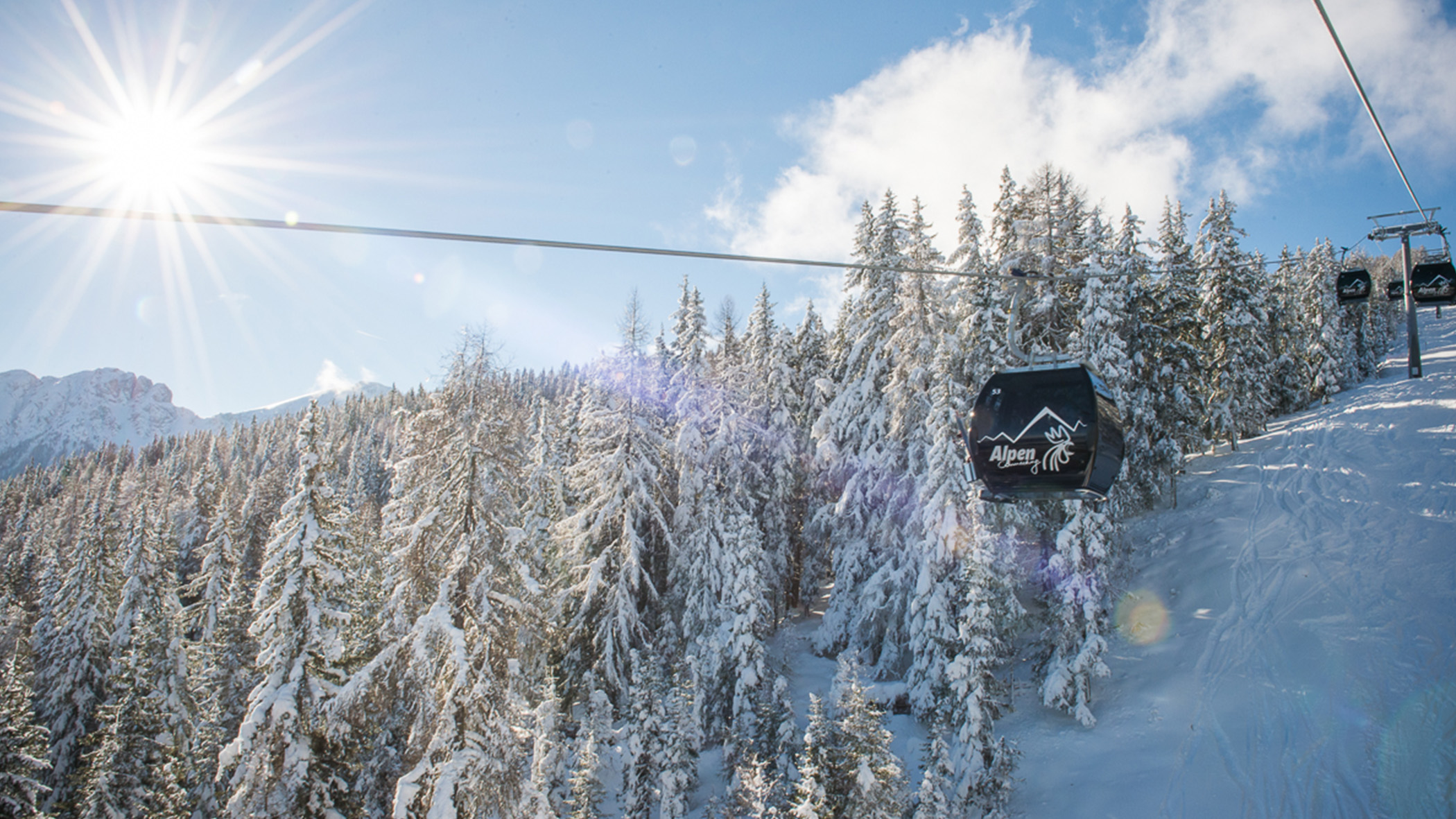 An einem sonnigen Wintertag in den Bergen gleiten schwarze Gondeln über verschneite Kiefern. - Mirabell Dolomites Hotel