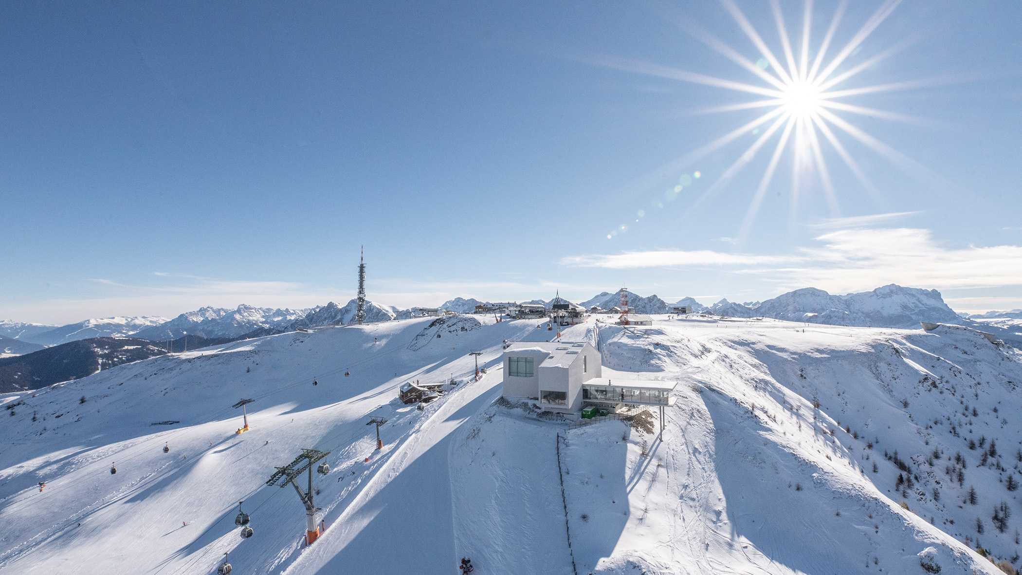 Ein verschneites Skigebiet in den Bergen unter strahlendem Sonnenschein, mit Sesselliften, Gebäuden und fernen Berggipfeln im Hintergrund. - Mirabell Dolomites Hotel