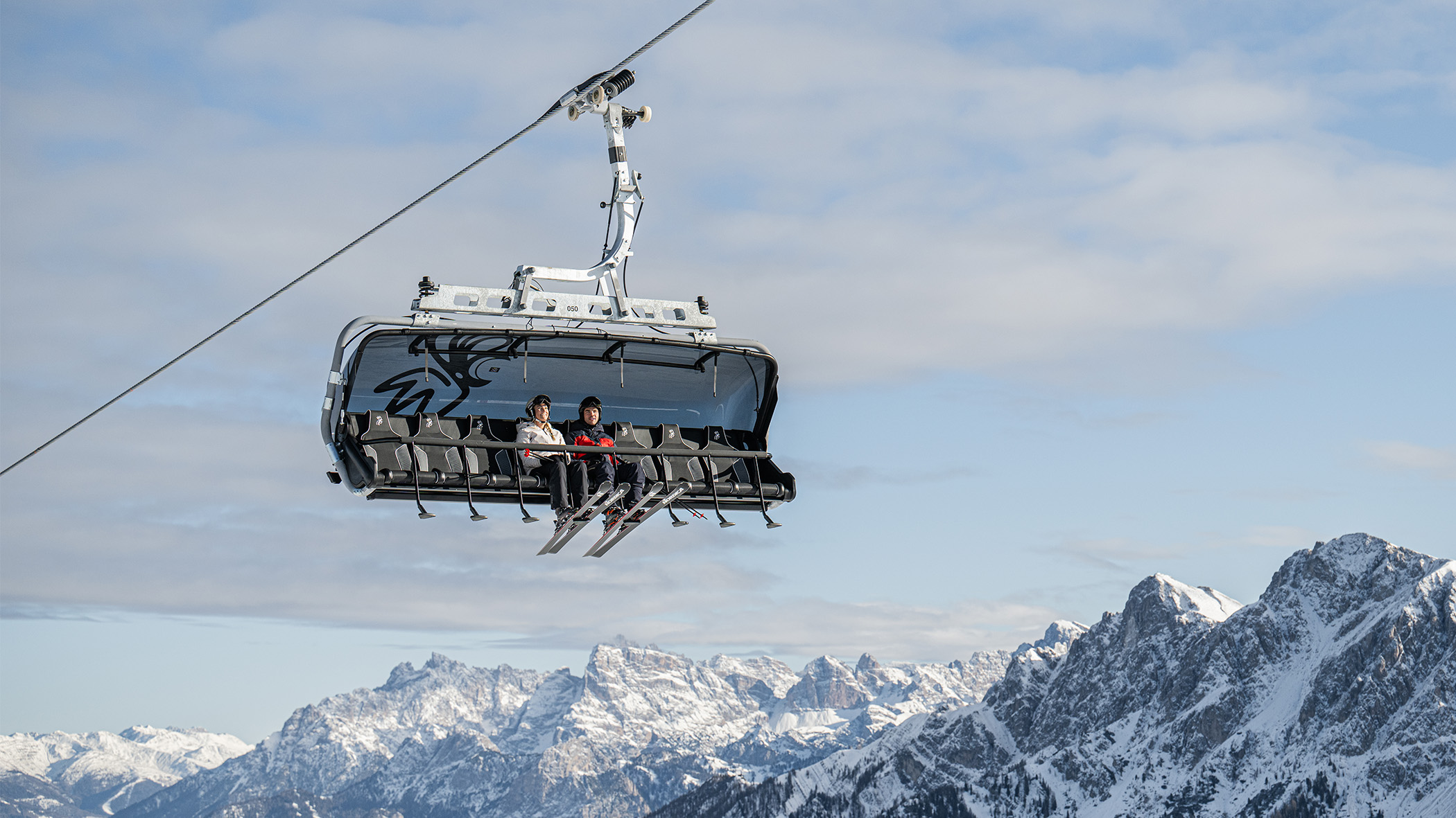 Drei Personen fahren mit einem Skilift über verschneite Berge hinweg, während der Himmel teilweise bewölkt ist. - Mirabell Dolomites Hotel