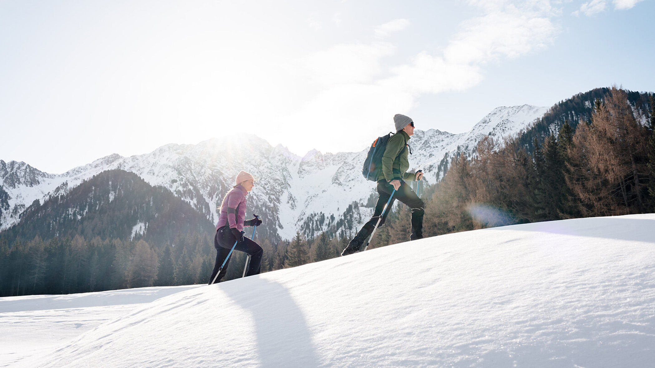 Zwei Menschen wandern bergauf durch den Schnee, im Hintergrund sind Bäume und Berge zu sehen, über ihnen erstreckt sich ein strahlender Himmel. - Mirabell Dolomites Hotel