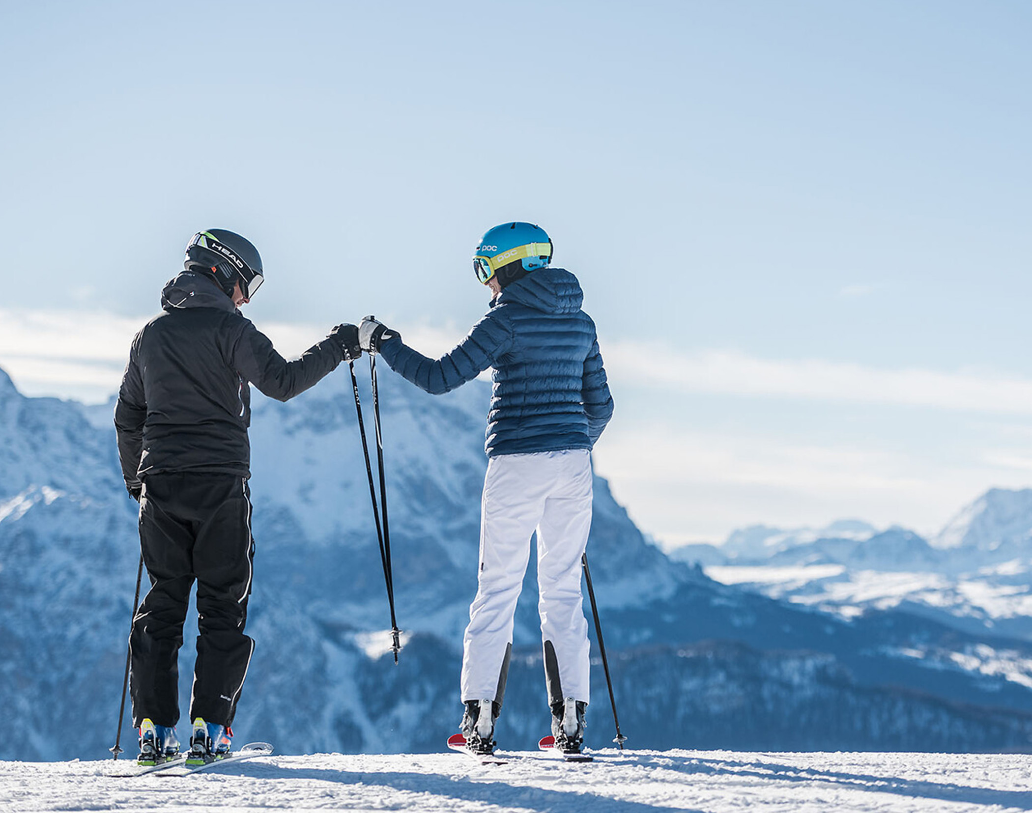 Zwei Skifahrer geben sich auf einem verschneiten Berg vor dem Hintergrund eines blauen Himmels und ferner Gipfel ein High Five. - Mirabell Dolomites Hotel