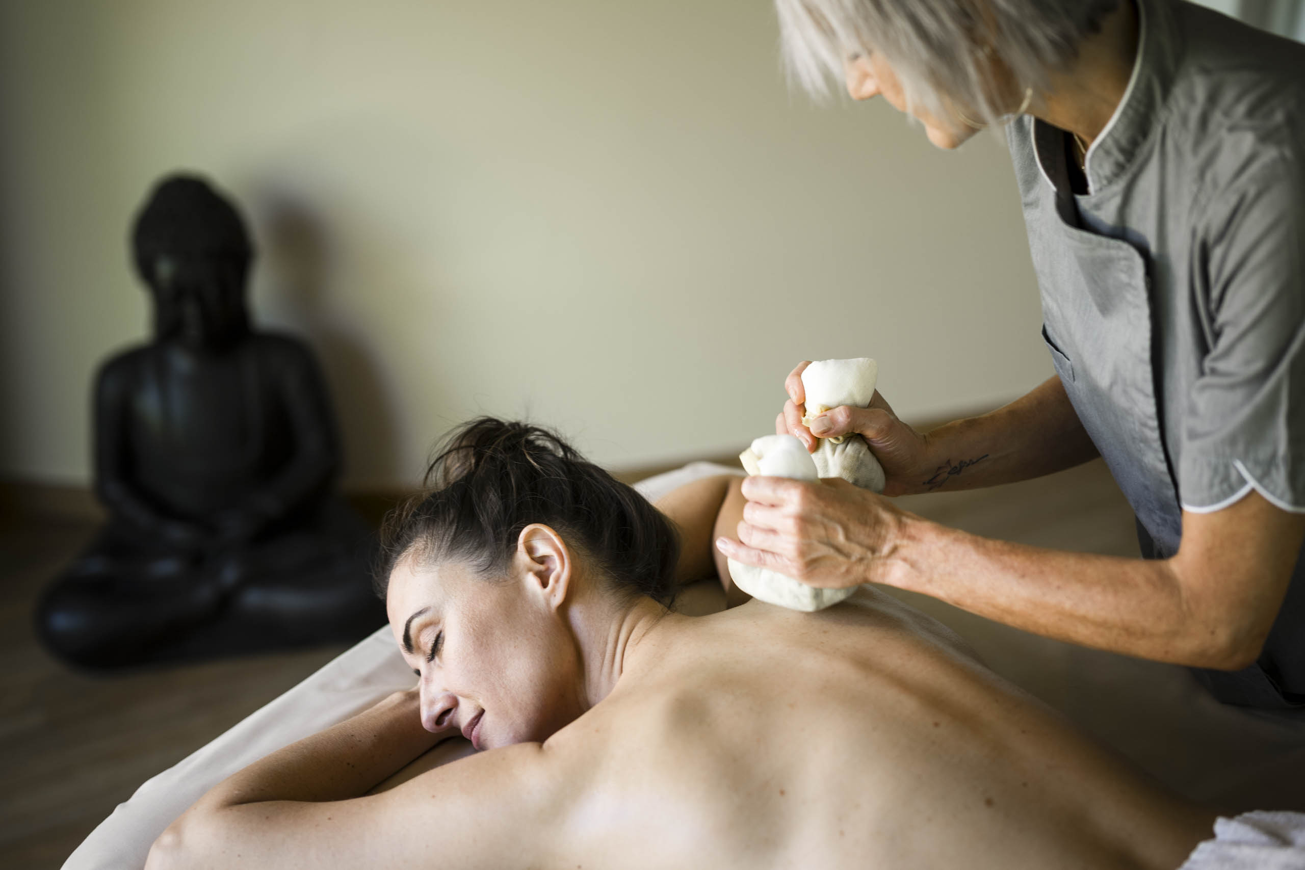 Eine Frau, die in einem Spa eine Kräuterkompressen-Massage am Rücken erhält, im Hintergrund eine Buddha-Statue. - Mirabell Dolomites Hotel