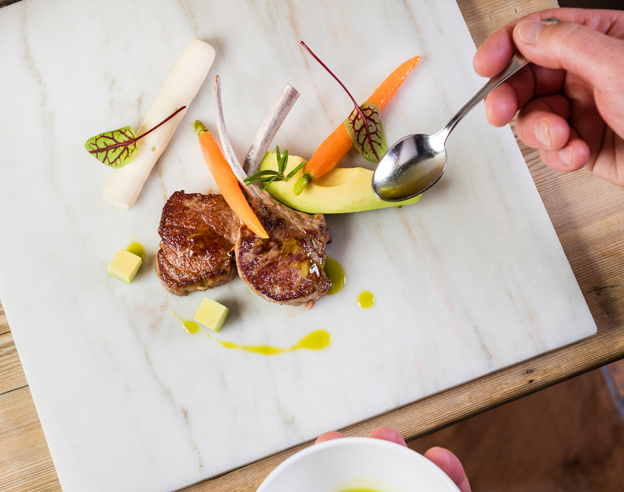 Eine Hand träufelt Soße auf ein Gourmetgericht mit Lamm, Möhren, Avocado und Kräutern, das auf einem Marmorteller angerichtet ist. - Mirabell Dolomites Hotel
