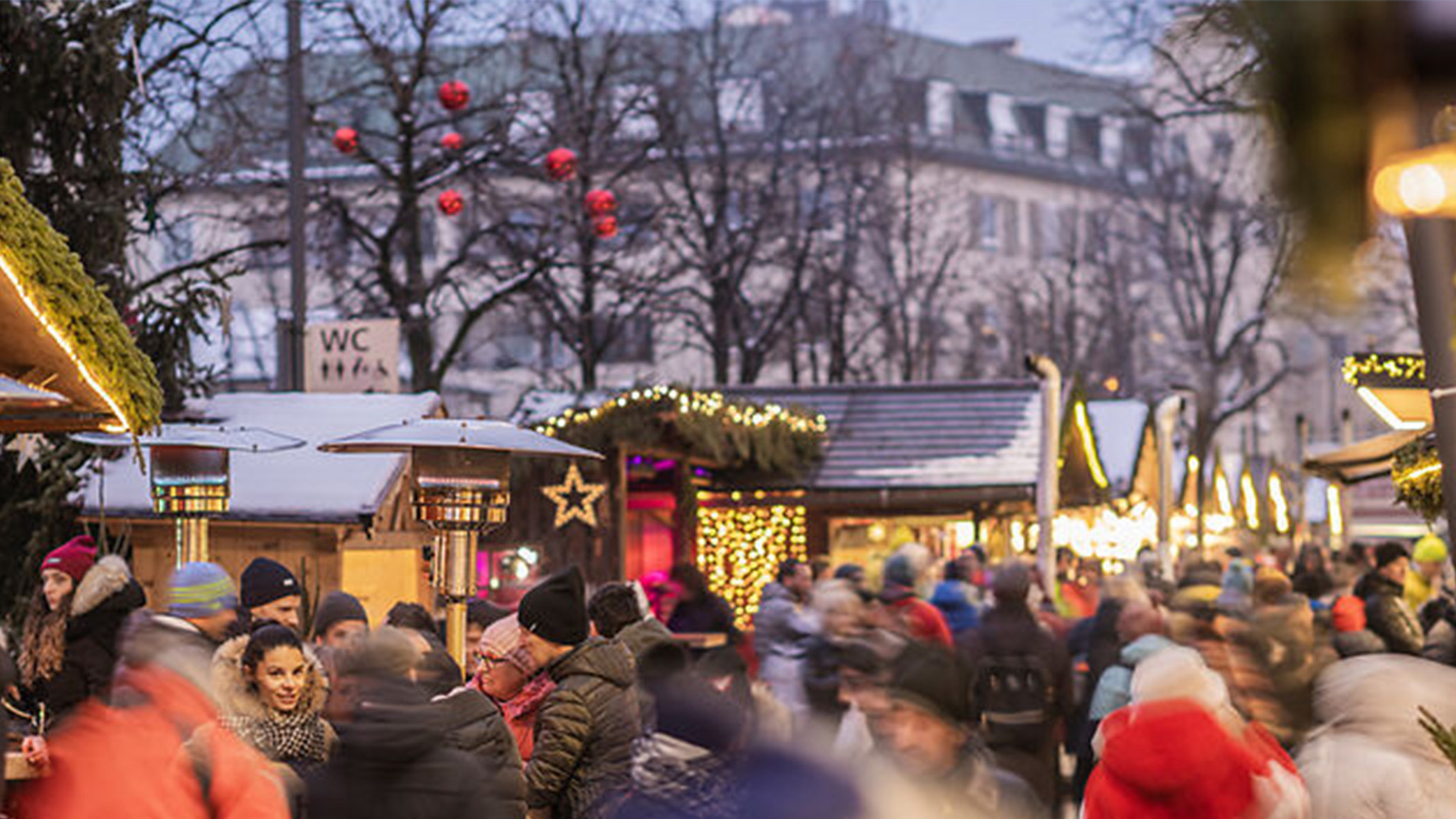 Die Menschen schlendern im Winter auf einem lebhaften Weihnachtsmarkt unter freiem Himmel an festlich geschmückten Ständen vorbei, umgeben von Lichtern und Bäumen. - Mirabell Dolomites Hotel
