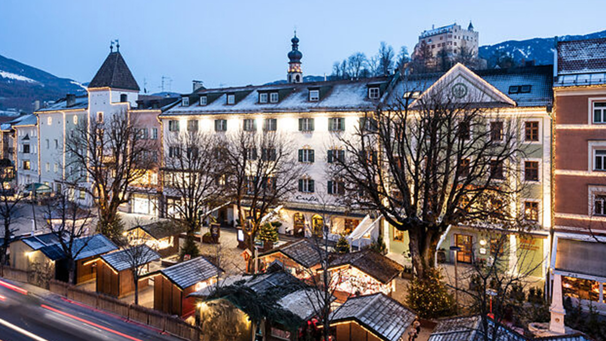 Ein festlich geschmückter europäischer Stadtplatz in der Abenddämmerung mit Lichtern, Marktständen und schneebedeckten Bergen im Hintergrund. - Mirabell Dolomites Hotel
