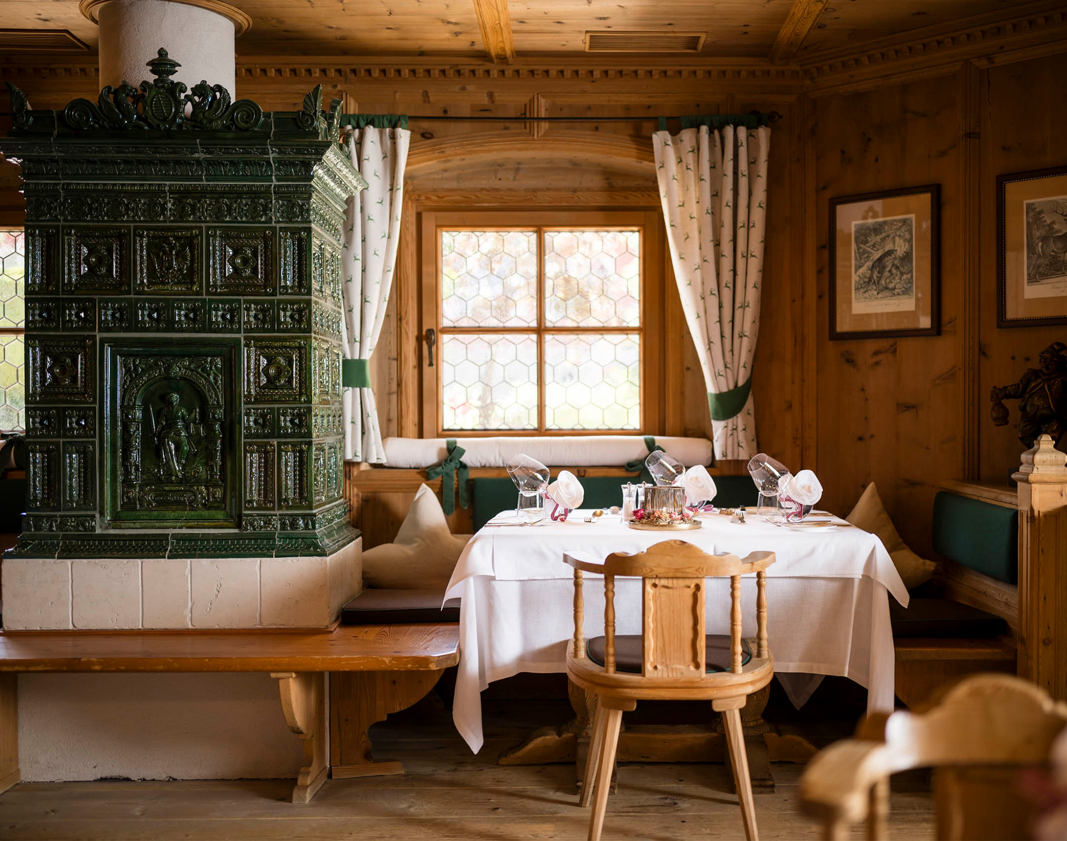 Gemütliches Esszimmer aus Holz mit einem grünen Kachelofen, einem gedeckten Tisch, einem Fenster und gerahmten Bildern an der Wand. - Mirabell Dolomites Hotel