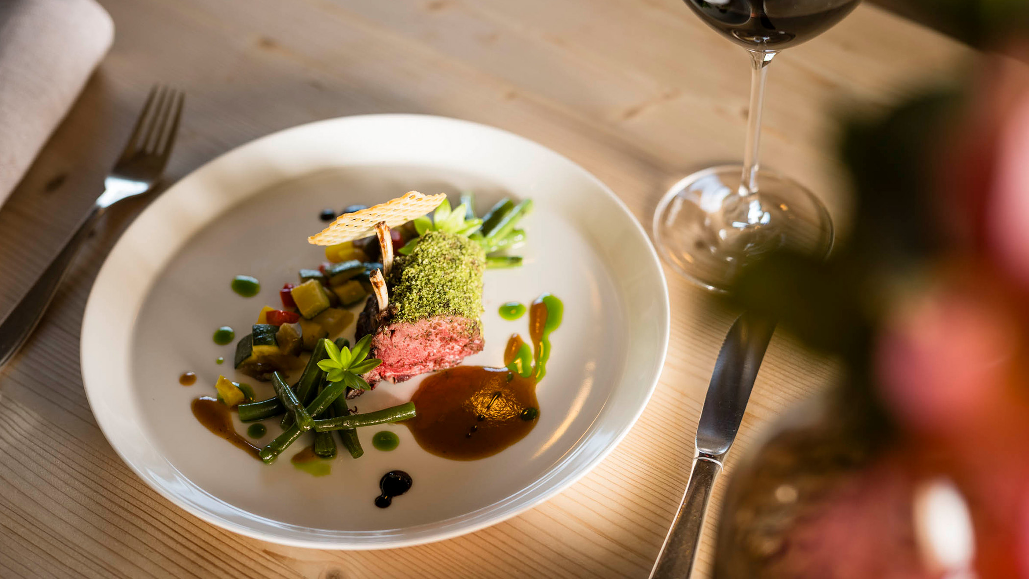 Ein Gourmetgericht mit Gemüse, Soße und Kräutern wird kunstvoll neben einem Glas Rotwein angerichtet. - Mirabell Dolomites Hotel