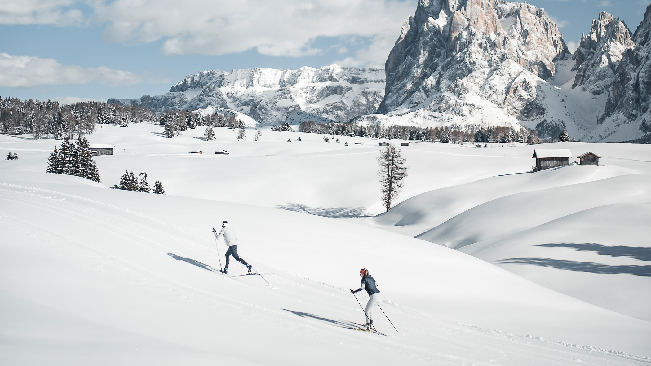 Zwei Menschen beim Langlaufen auf verschneiten Hügeln, im Hintergrund Berge und Bäume. - Mirabell Dolomites Hotel