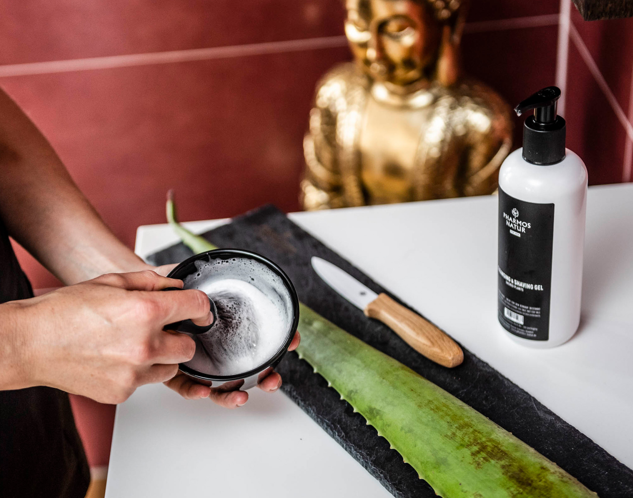 Seife, die in einer Schüssel von Hand aufgeschäumt wird, ein Aloe-Vera-Blatt, ein Messer und eine Lotionsflasche auf einem Tisch, im Hintergrund eine goldene Statue. - Mirabell Dolomites Hotel