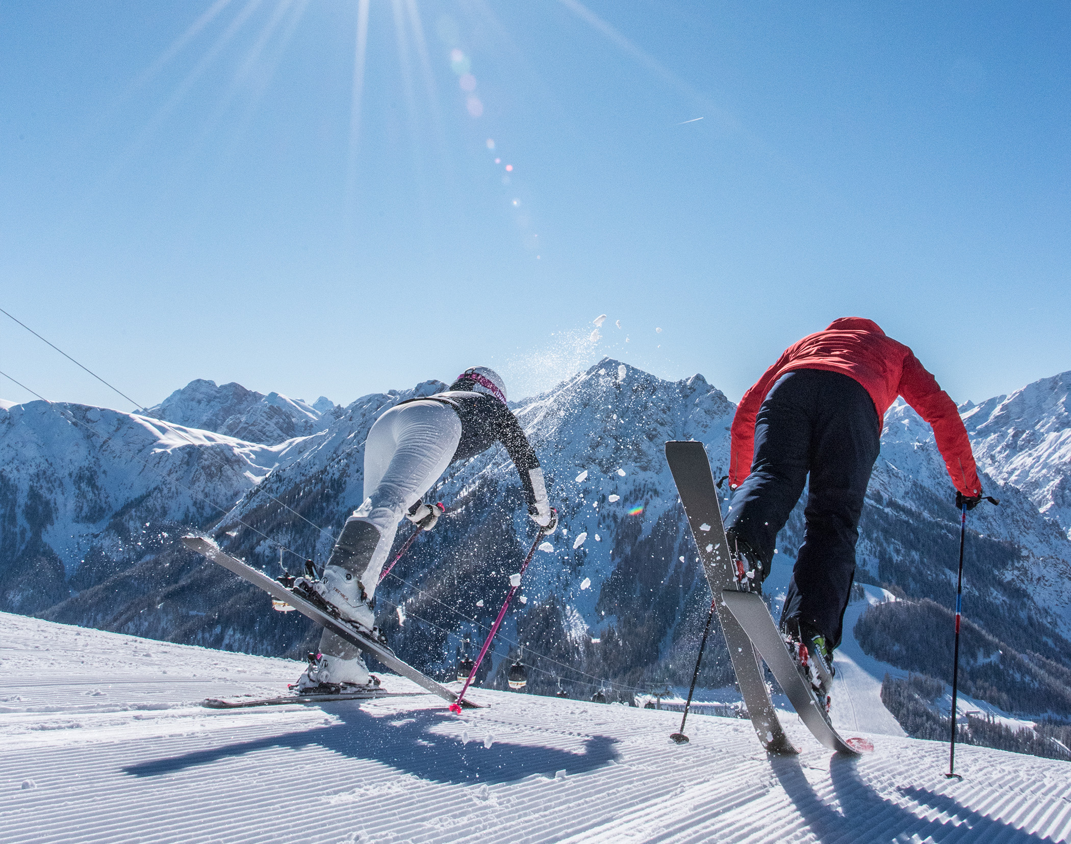 Zwei Skifahrer in Bewegung auf einem verschneiten Hang, im Hintergrund Berge und ein strahlend blauer Himmel. - Mirabell Dolomites Hotel